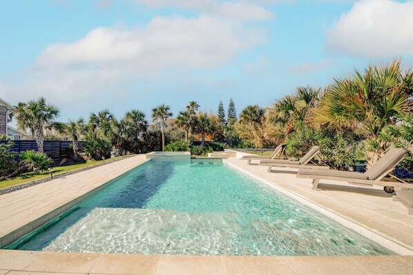 Piscina rectangular con agua cristalina, rodeada de palmeras y sillas de descanso.