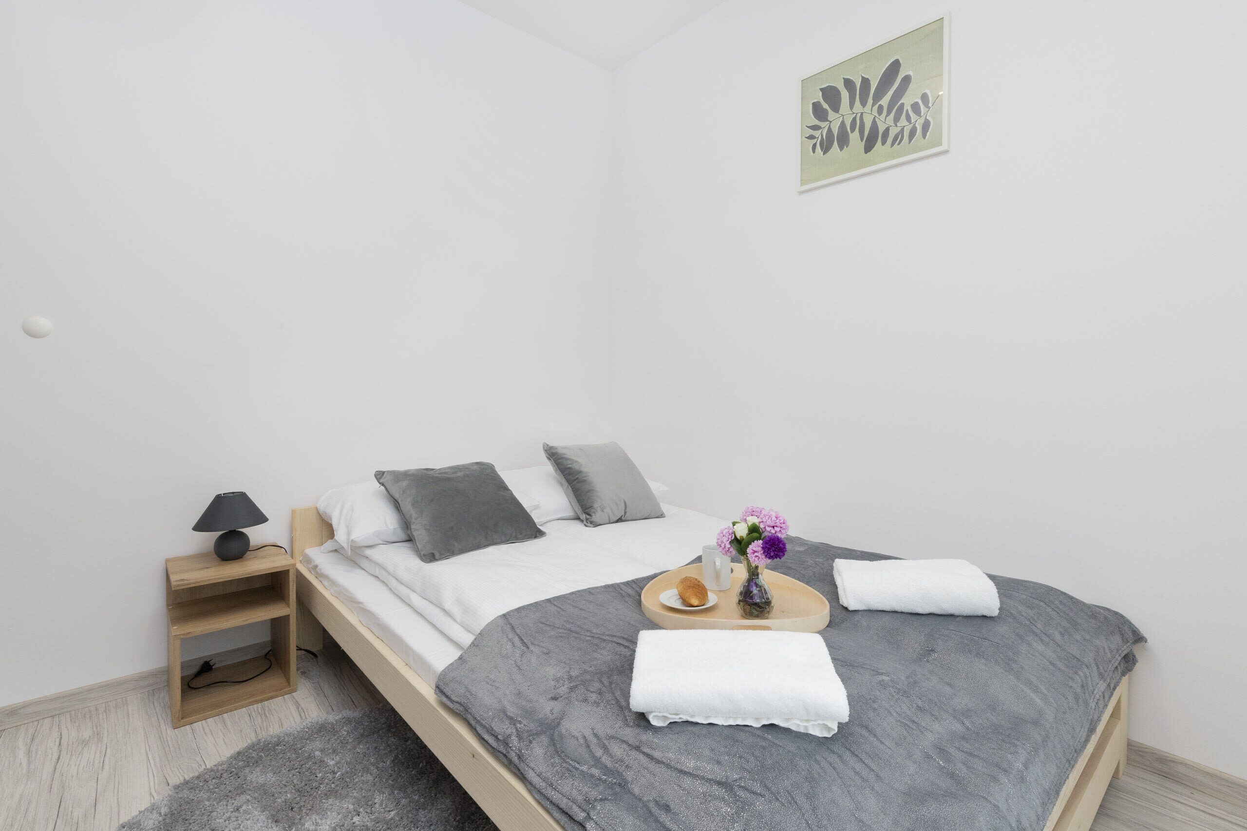 A neatly made bed with gray bedding, a bedside table with a lamp, and a tray with a muffin, a glass of water, and a vase of flowers.