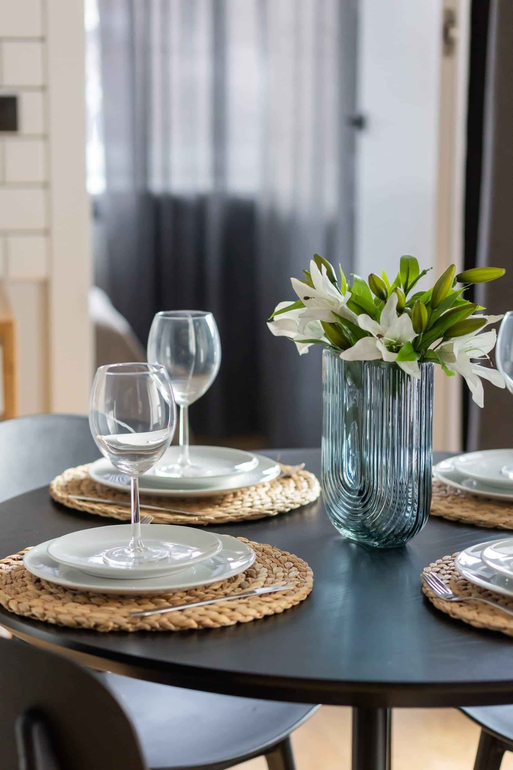 A dining table with a glass vase containing white flowers, two wine glasses, and plates on woven placemats.