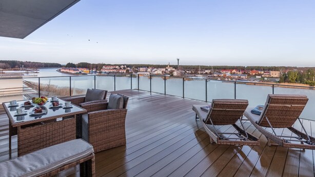 A deck with wicker furniture, a table with plates and a fruit bowl, overlooking a river with buildings in the distance. - Spacious Apartment | Terrace | Parking| SPA | Pool (Dziwnów)