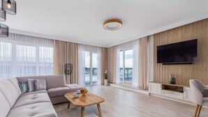 A modern living room with a sofa, coffee table, TV, and large windows. - Spacious Apartment | Terrace | Parking| SPA | Pool (Dziwnów)
