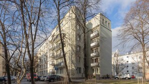Un immeuble résidentiel à plusieurs étages, doté de balcons, avec des voitures garées et des arbres, situé dans un quartier urbain ensoleillé.