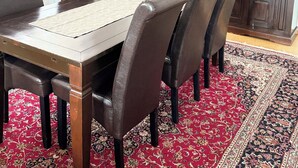 Ein Essbereich mit einem Holztisch, Ledersesseln, einem Teppich mit rotem Muster und einer Küche im Hintergrund.