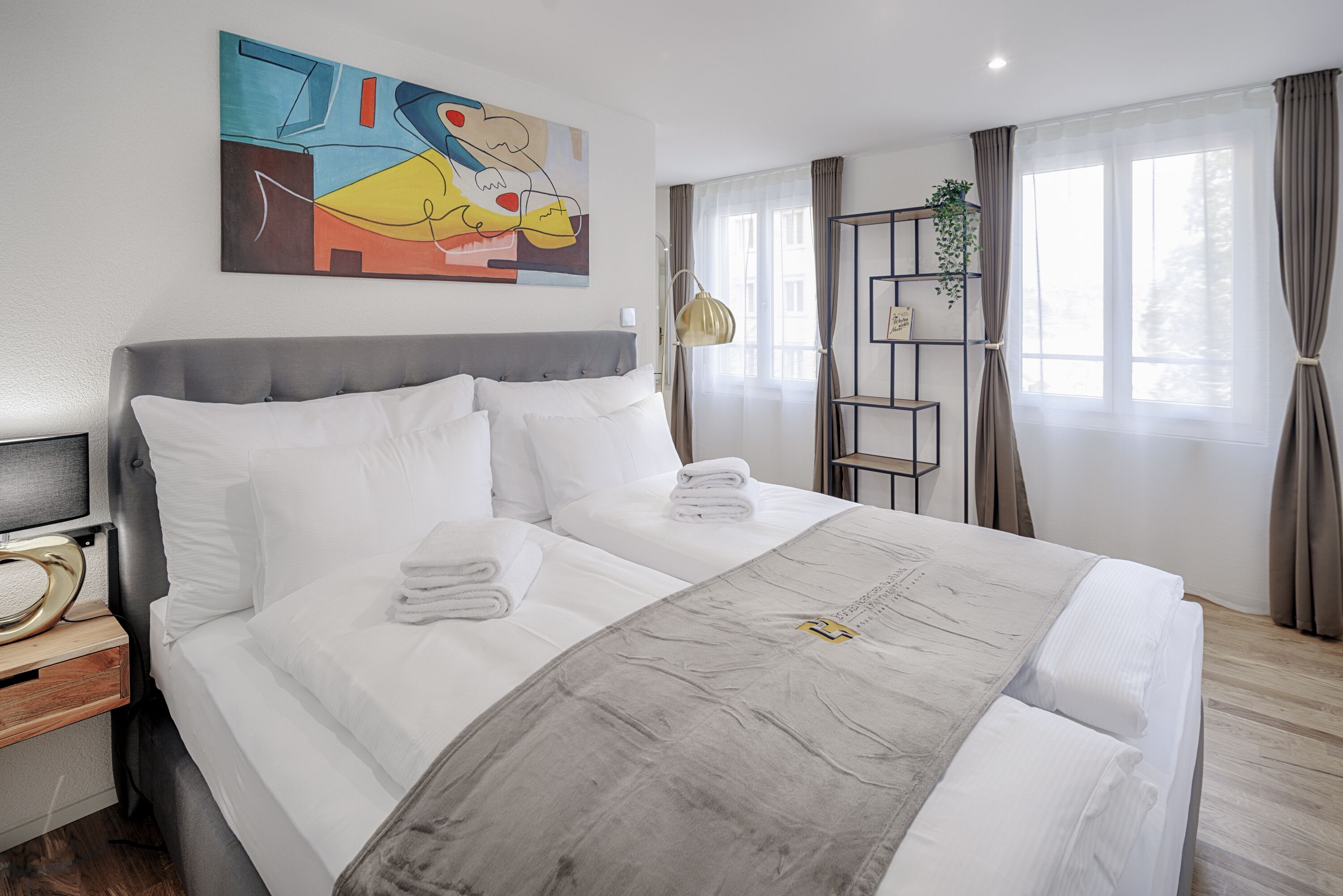 A hotel room with a large bed, a bedside table with a lamp, a painting on the wall, and a window with curtains.