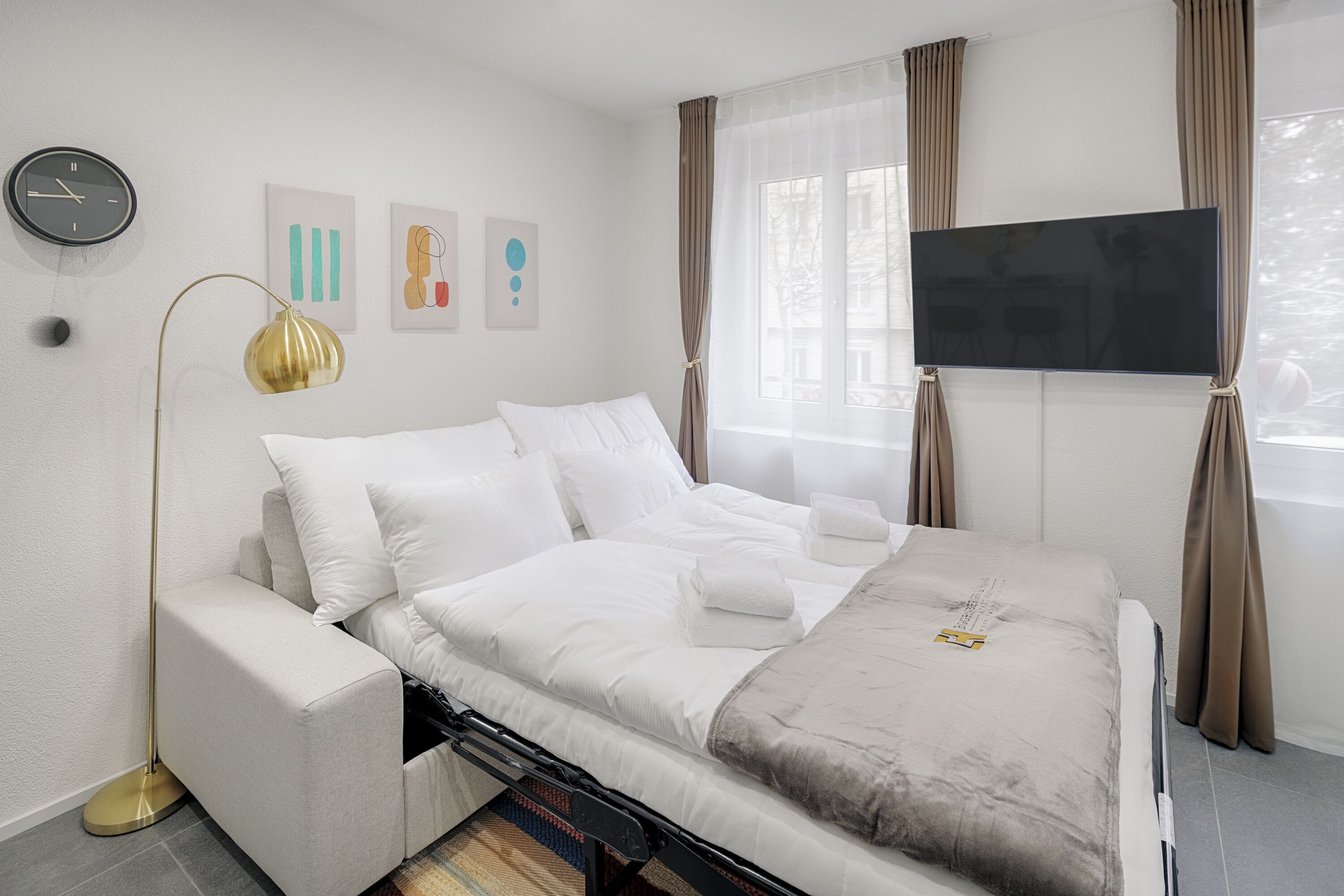 Una habitación de hotel moderna con una cama, un televisor de pantalla plana, un reloj de pared y obras de arte en la pared.