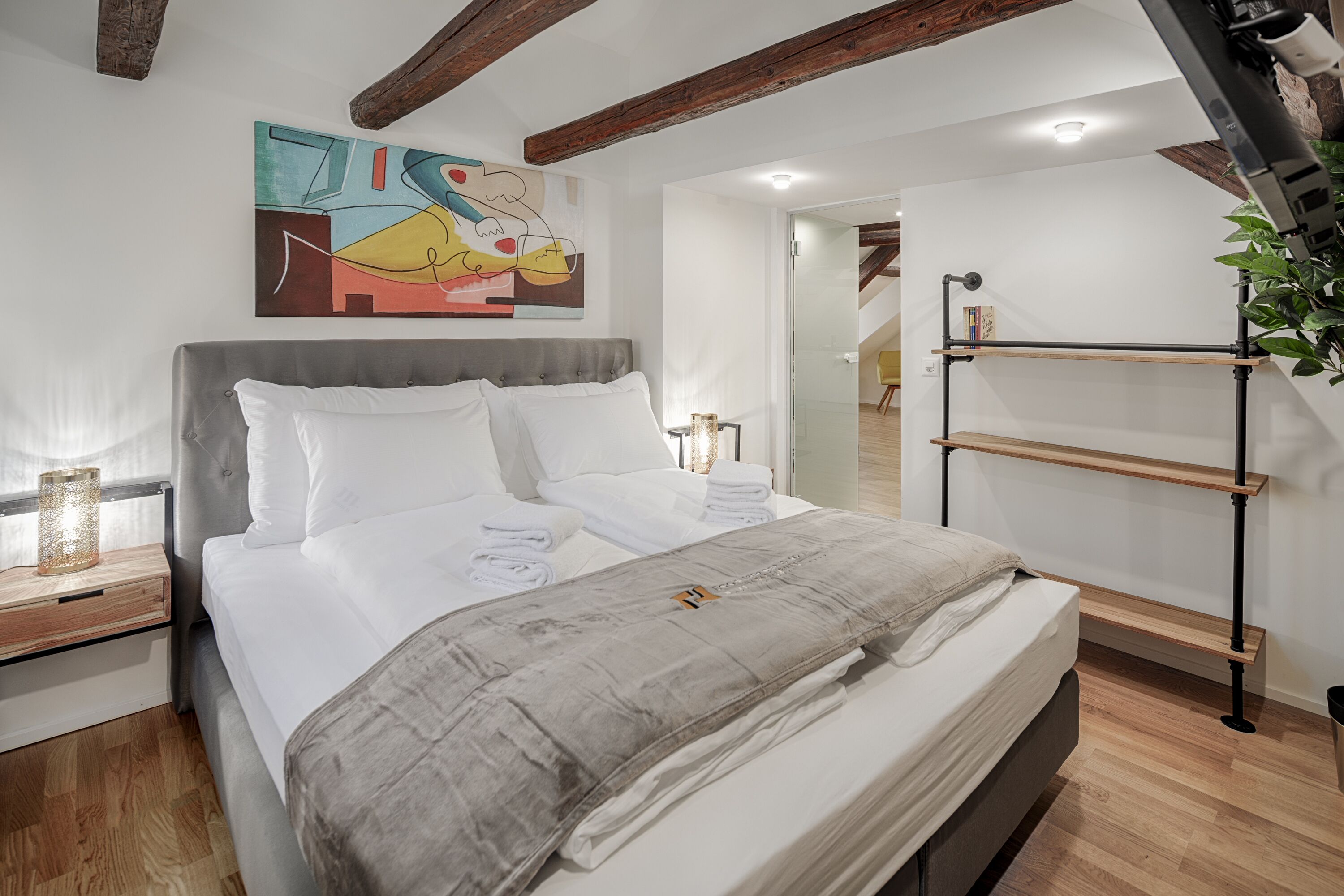 A modern bedroom with a large bed, a wooden headboard, a painting on the wall, and a loft-style staircase.