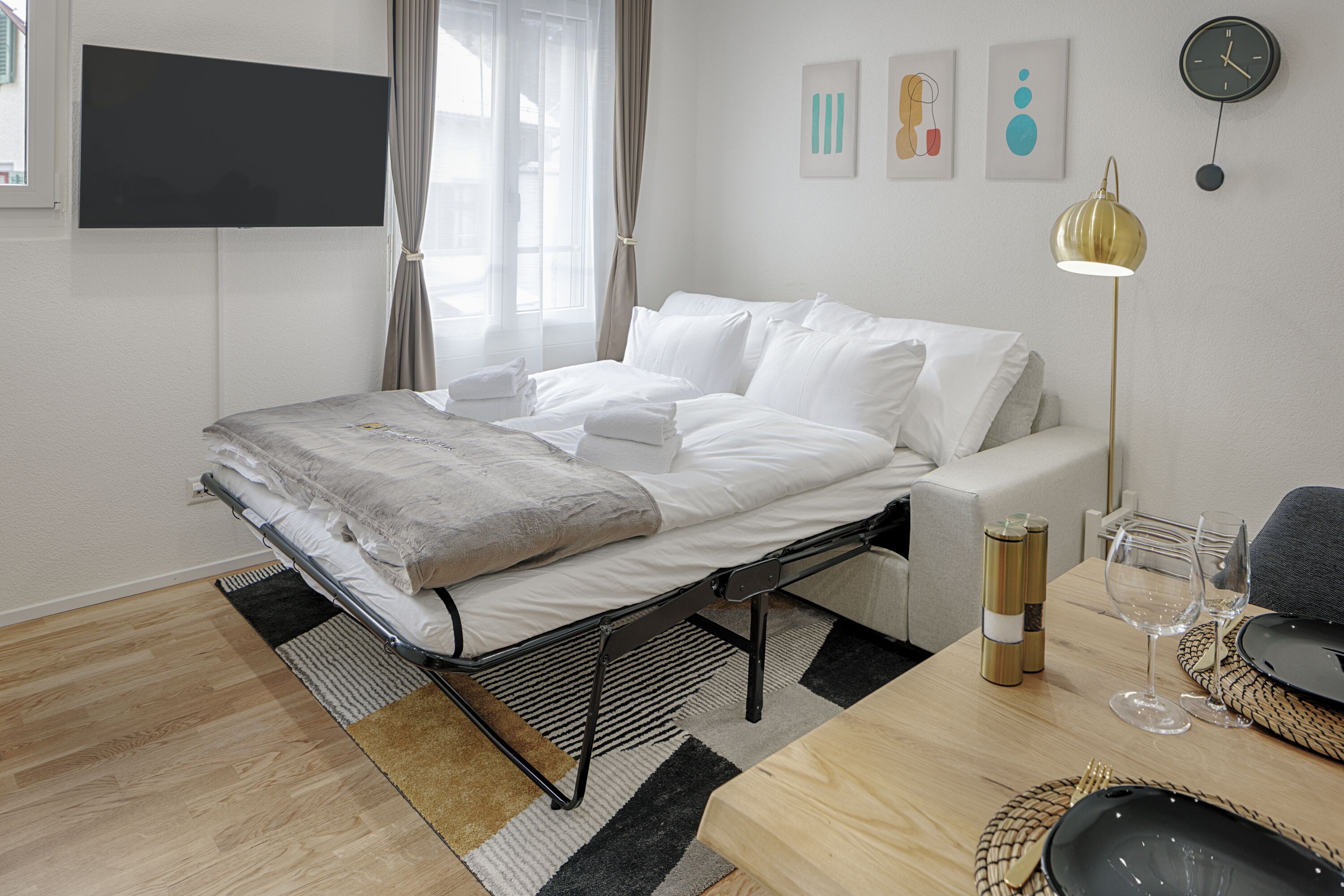 A modern hotel room with a bed, a dining table set for two, a flat-screen TV mounted on the wall, and a clock.