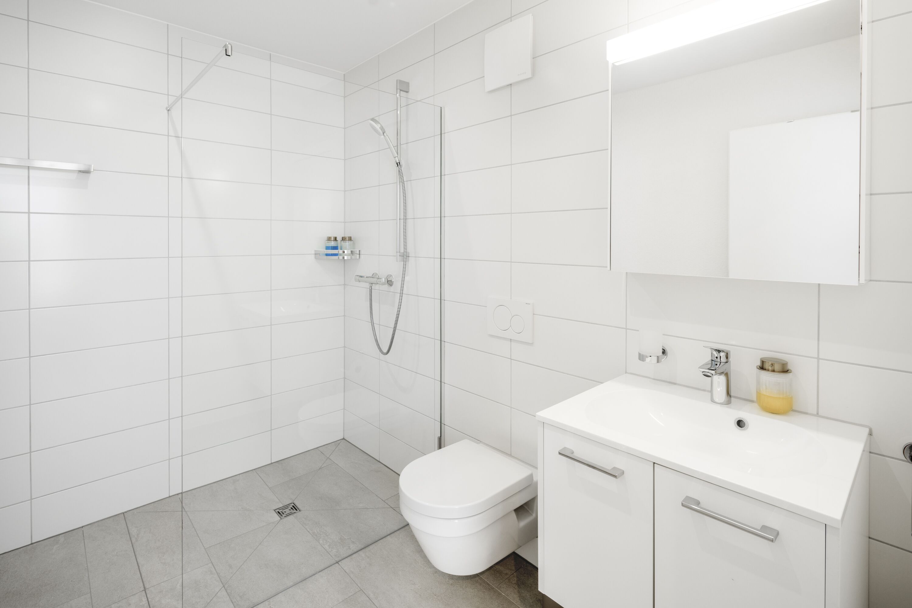 Egy modern fürőszoba fehér csempékkel, zuhanyzóval, WC-vel és egy mosdókagylóval, mely felett tükör található.
