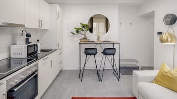 Una cocina moderna con armarios blancos, encimera negra y horno integrado. Hay un comedor con un espejo redondo, dos sillas negras y una pequeña mesa. Se ve un sofá blanco con un cojín amarillo en primer plano.