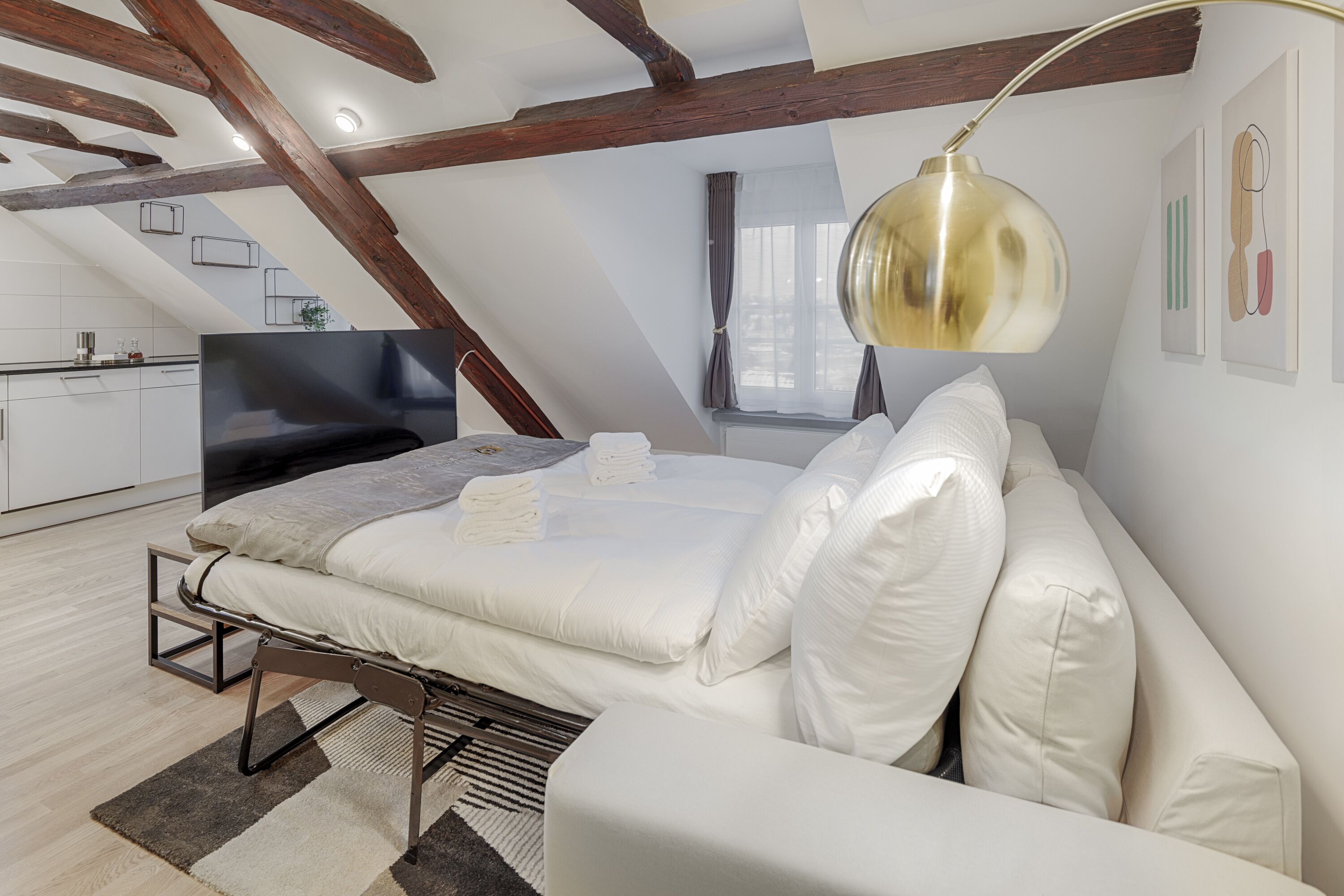 A modern loft bedroom with a large bed, a TV, and a dining area visible through an open door.