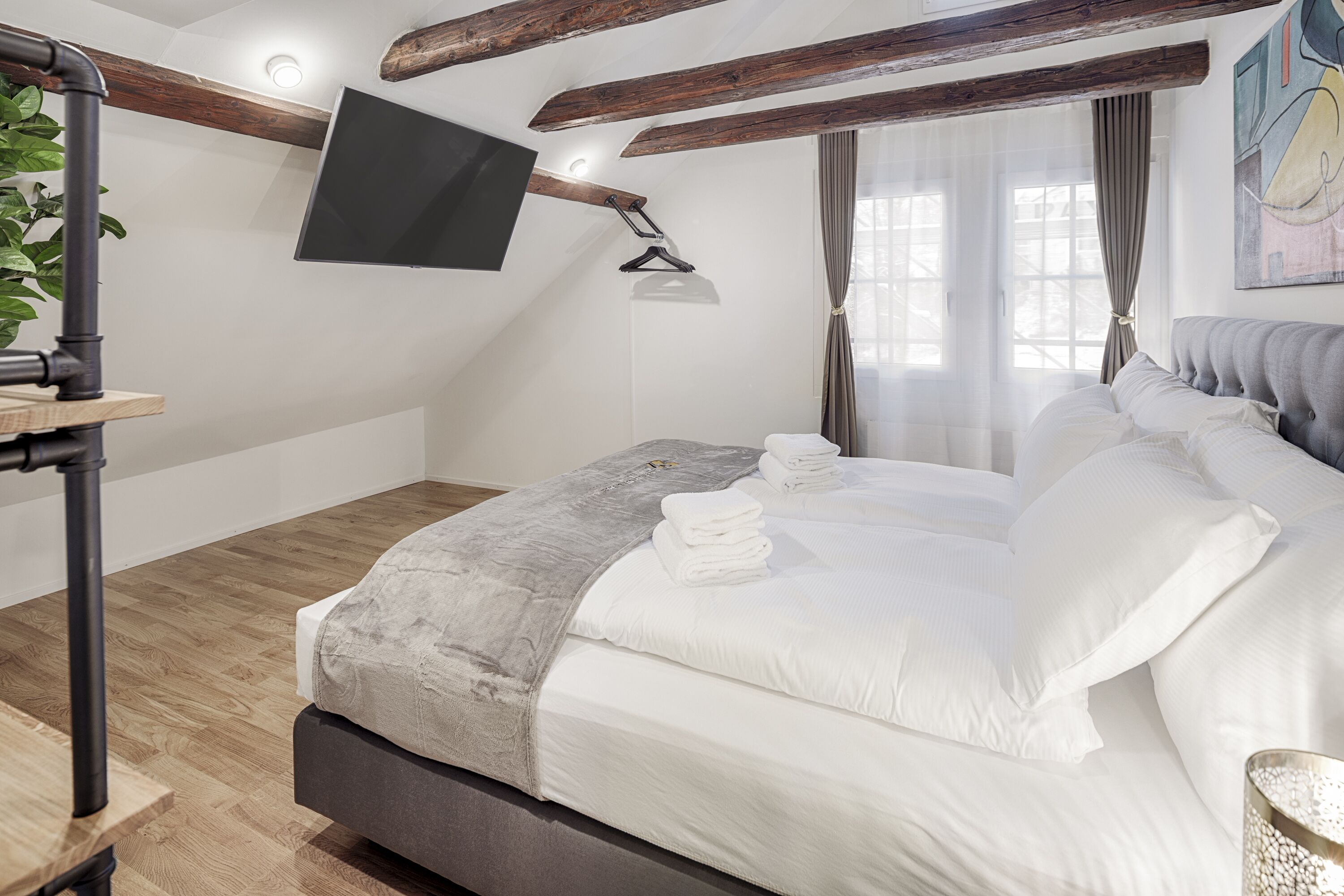Une chambre à coucher avec un lit, un téléviseur fixé au mur, un plancher en bois et une fenêtre avec des rideaux.