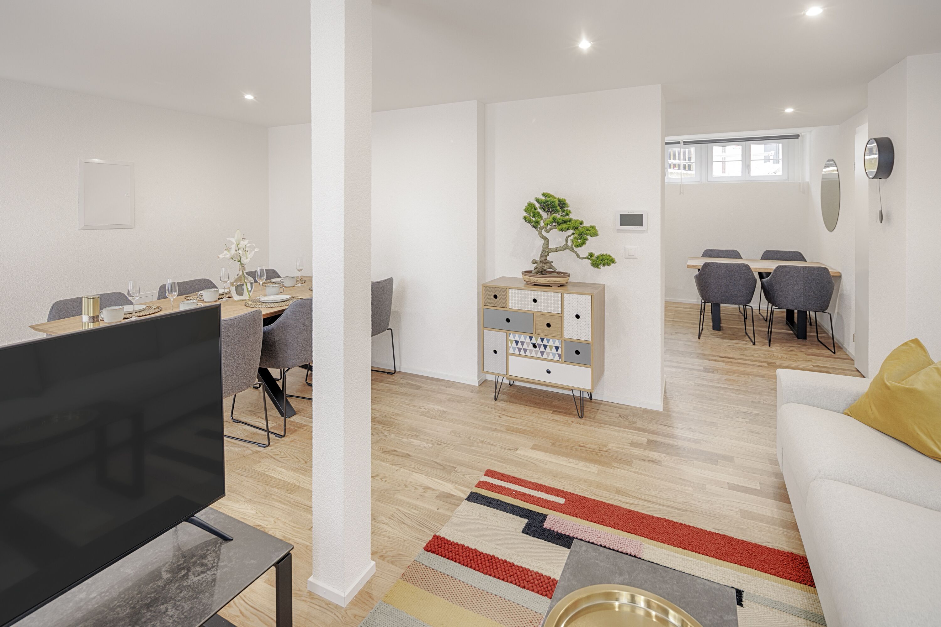 Un salon moderne doté d’un téléviseur à écran plat, d’un canapé, d’un coin repas avec une table dressée pour quatre personnes et d’une petite armoire contenant un bonsaï.
