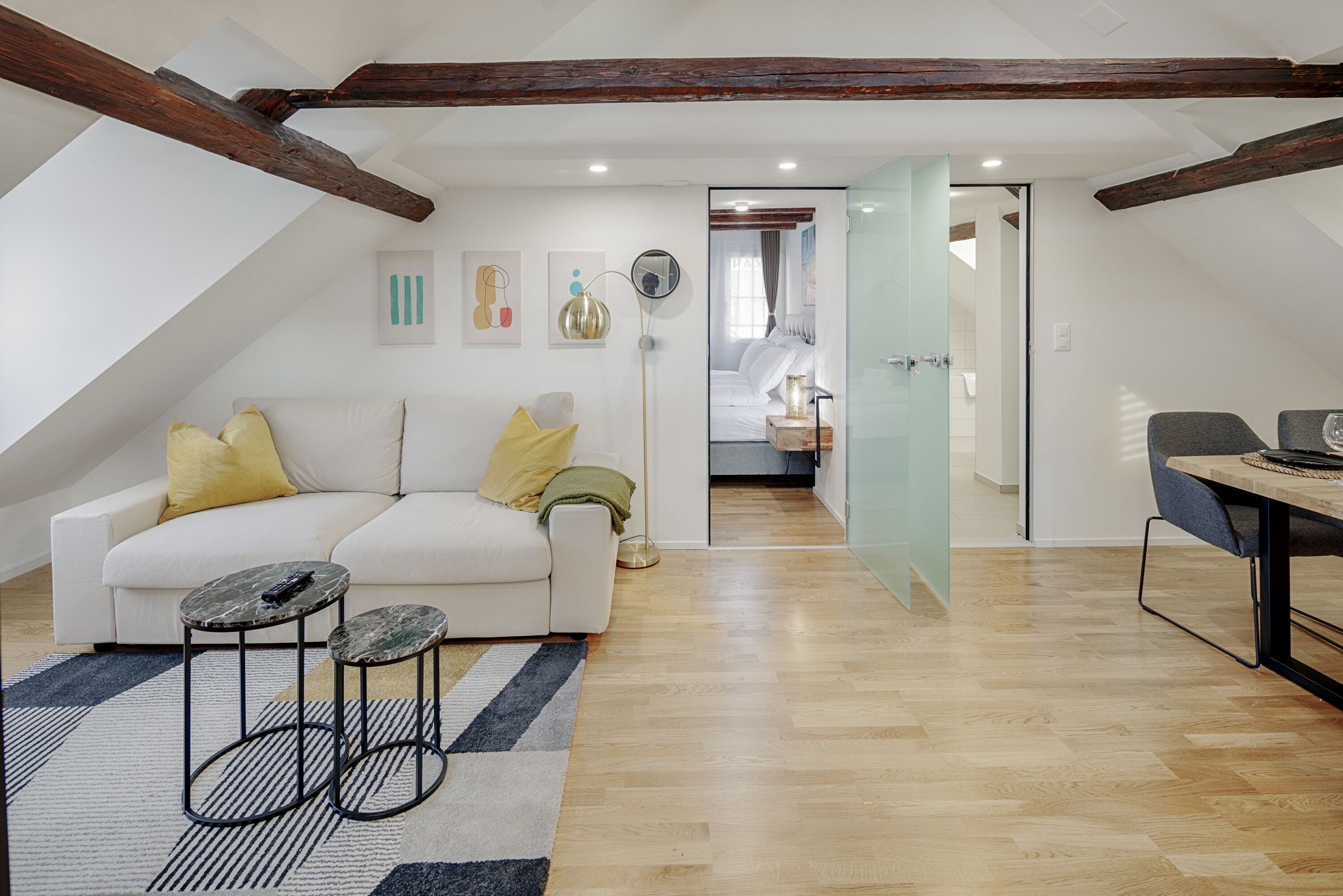 Ein modernes Schlafzimmer im Loft-Stil mit schrägverdachter Decke, einem weißen Sofa mit gelben Kissen, einem kleinen runden Tisch und einer Glastür zum Badezimmer.