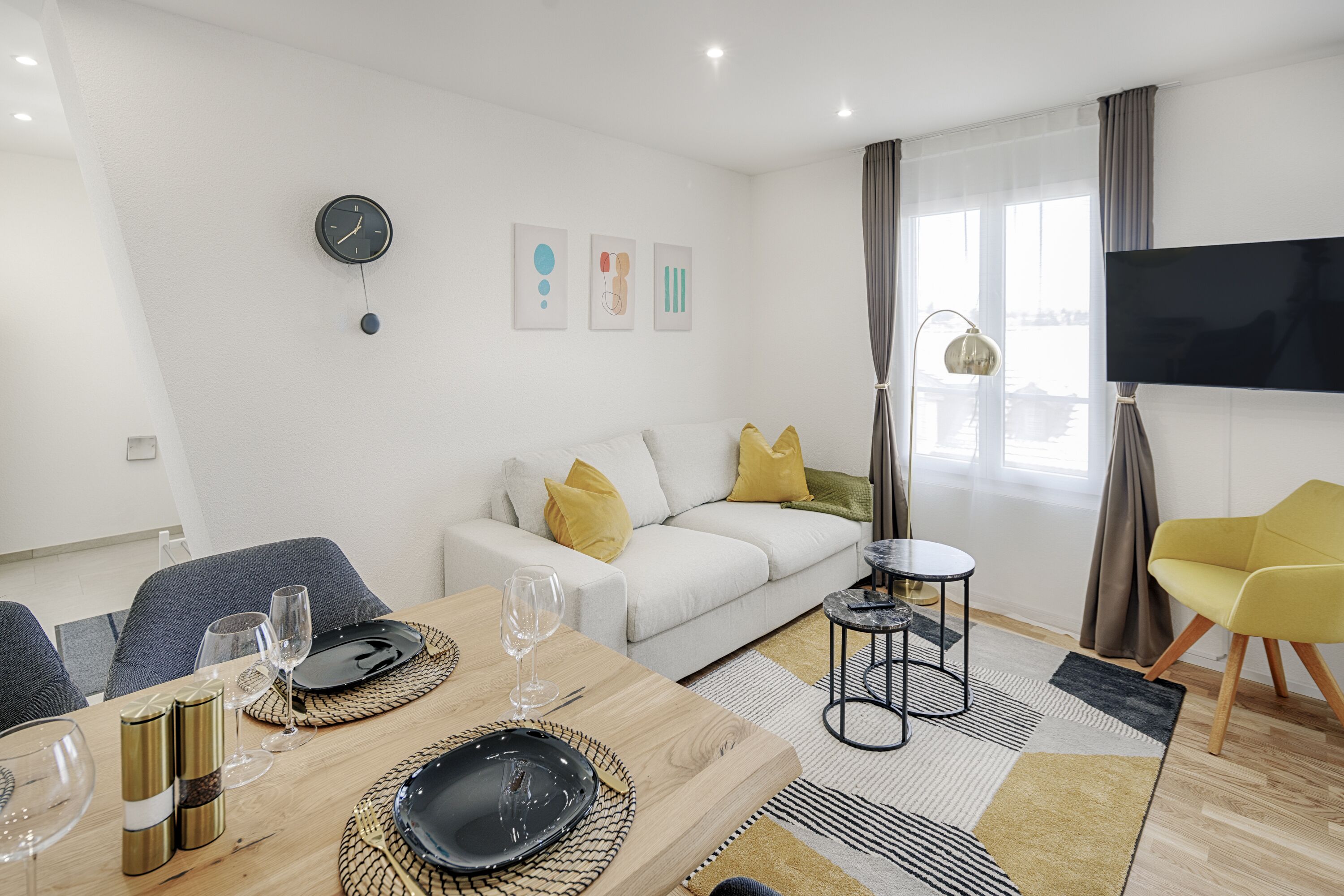 Un salon moderne comprenant un coin repas, un canapé avec des coussins jaunes, un téléviseur à écran plat et une grande fenêtre avec des rideaux.