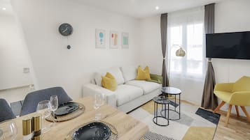 Un salon moderne comprenant un coin repas, un canapé avec des coussins jaunes, un téléviseur à écran plat et une grande fenêtre avec des rideaux.