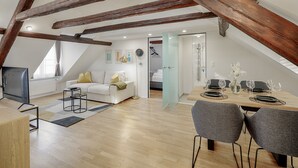 Un loft moderno con vigas de madera a la vista, un comedor con separador de vidrio, una sala de estar con sofá y televisor, y una cocina al fondo.