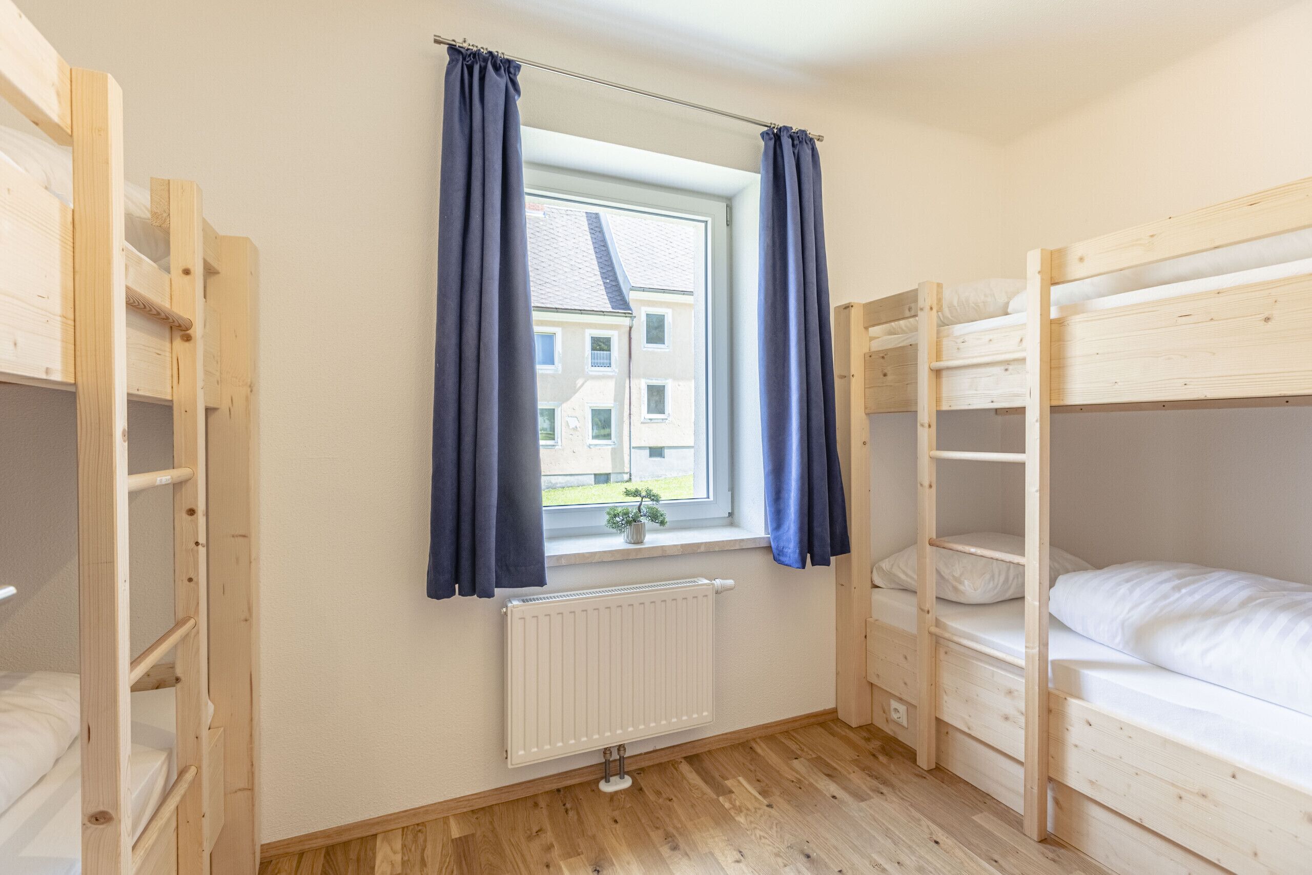 A dormitory room with bunk beds, a window with curtains, a radiator, and a view of a building outside.