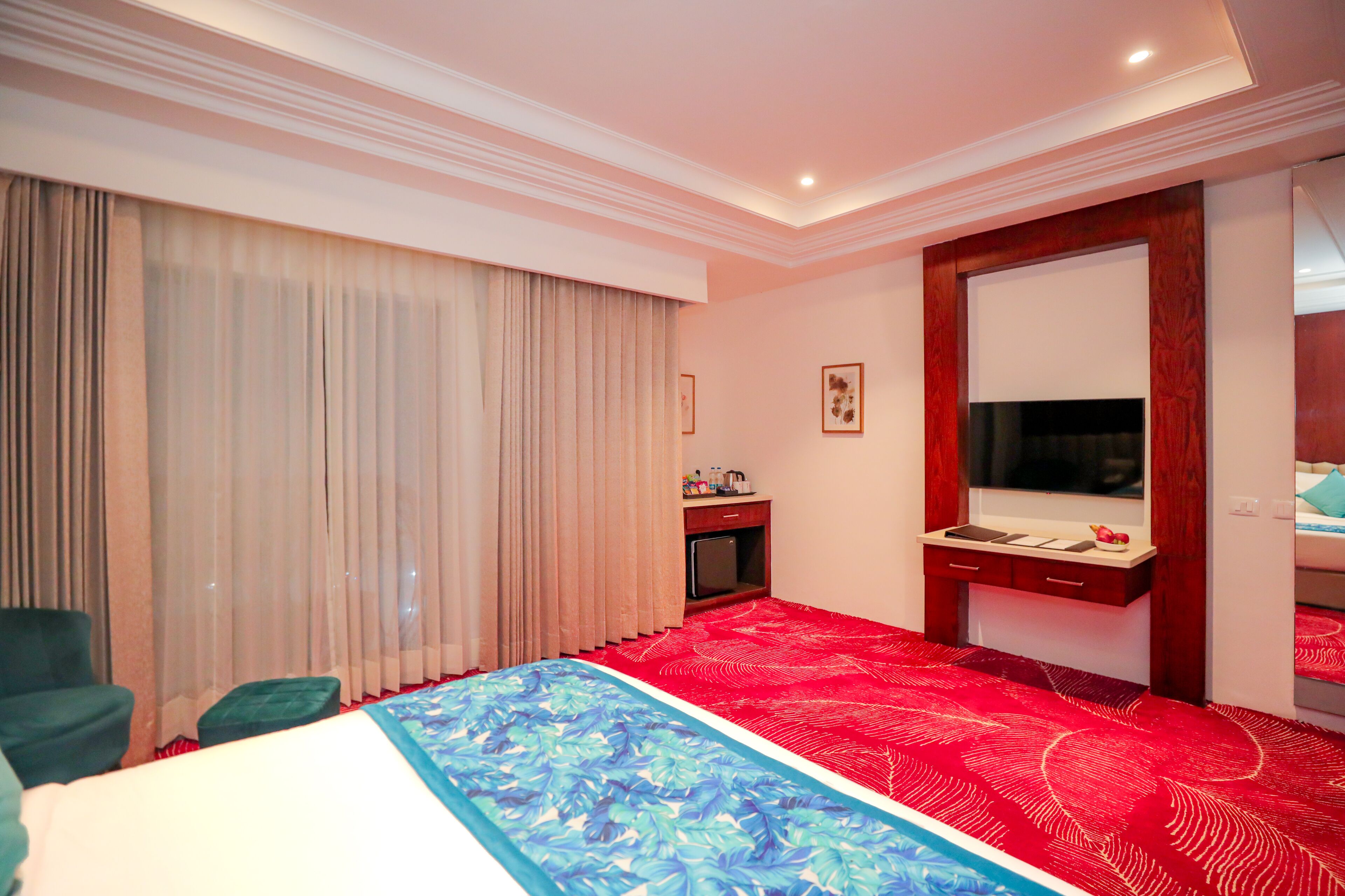 Ein Hotelzimmer mit einem großen Bett, einem Schreibtisch mit Spiegel, einem Fernseher und einem Fenster mit Vorhängen.