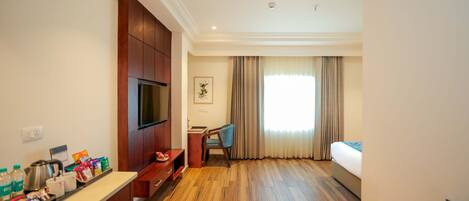 A hotel room with a wooden floor, a bed, a TV, a desk with a chair, and a window with curtains.