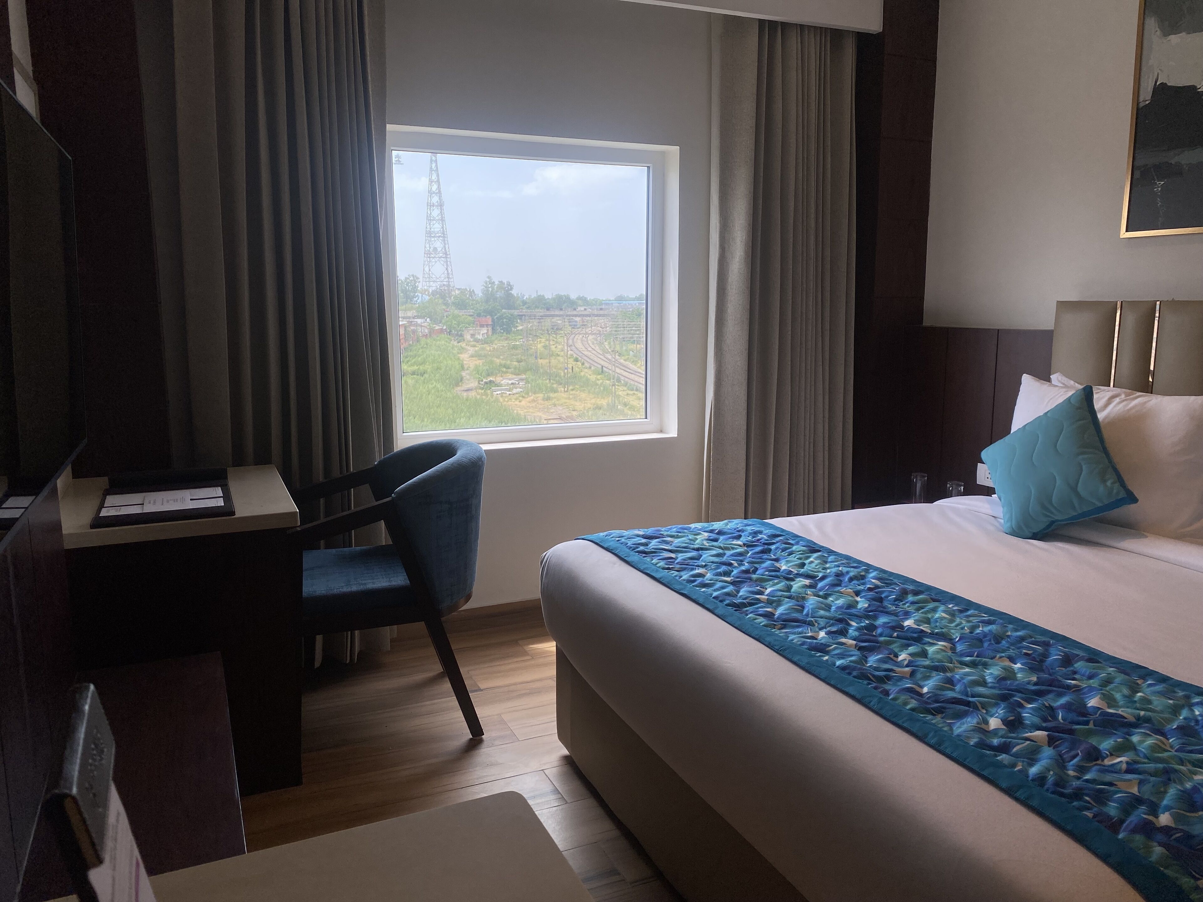 Habitación de hotel con una cama grande, un escritorio con una silla, un ventanal con vistas a una vía ferroviaria y una cama con almohada azul y blanca.