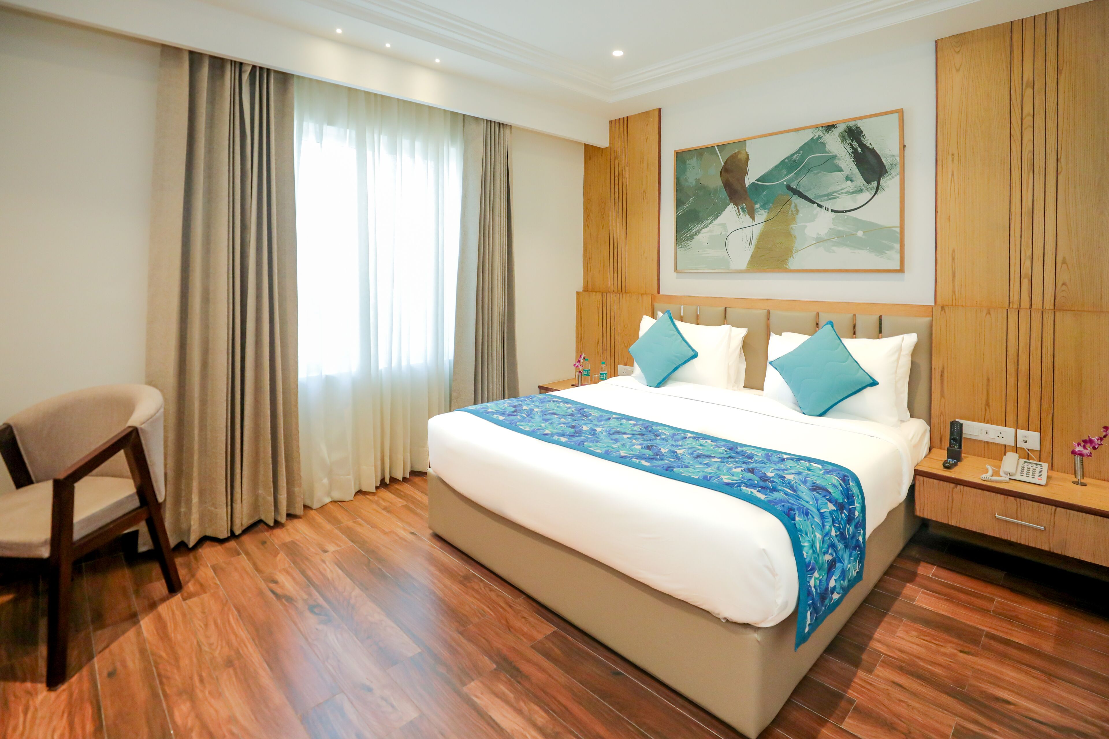 A hotel room with a large bed, wooden flooring, a chair, a nightstand with a phone, and a painting on the wall.