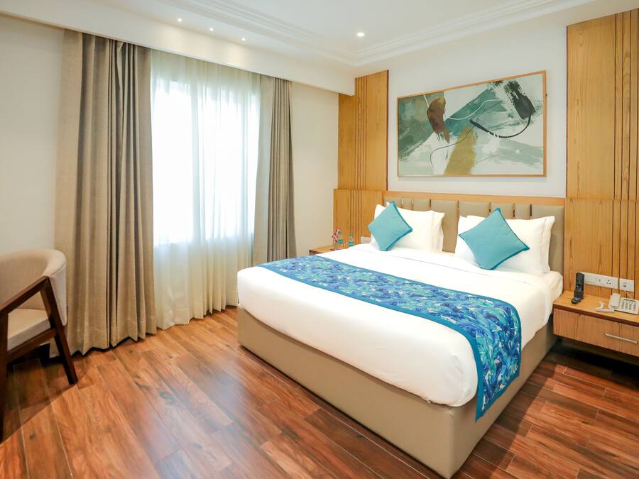 A hotel room with a large bed, wooden flooring, a chair, a nightstand with a phone, and a painting on the wall.