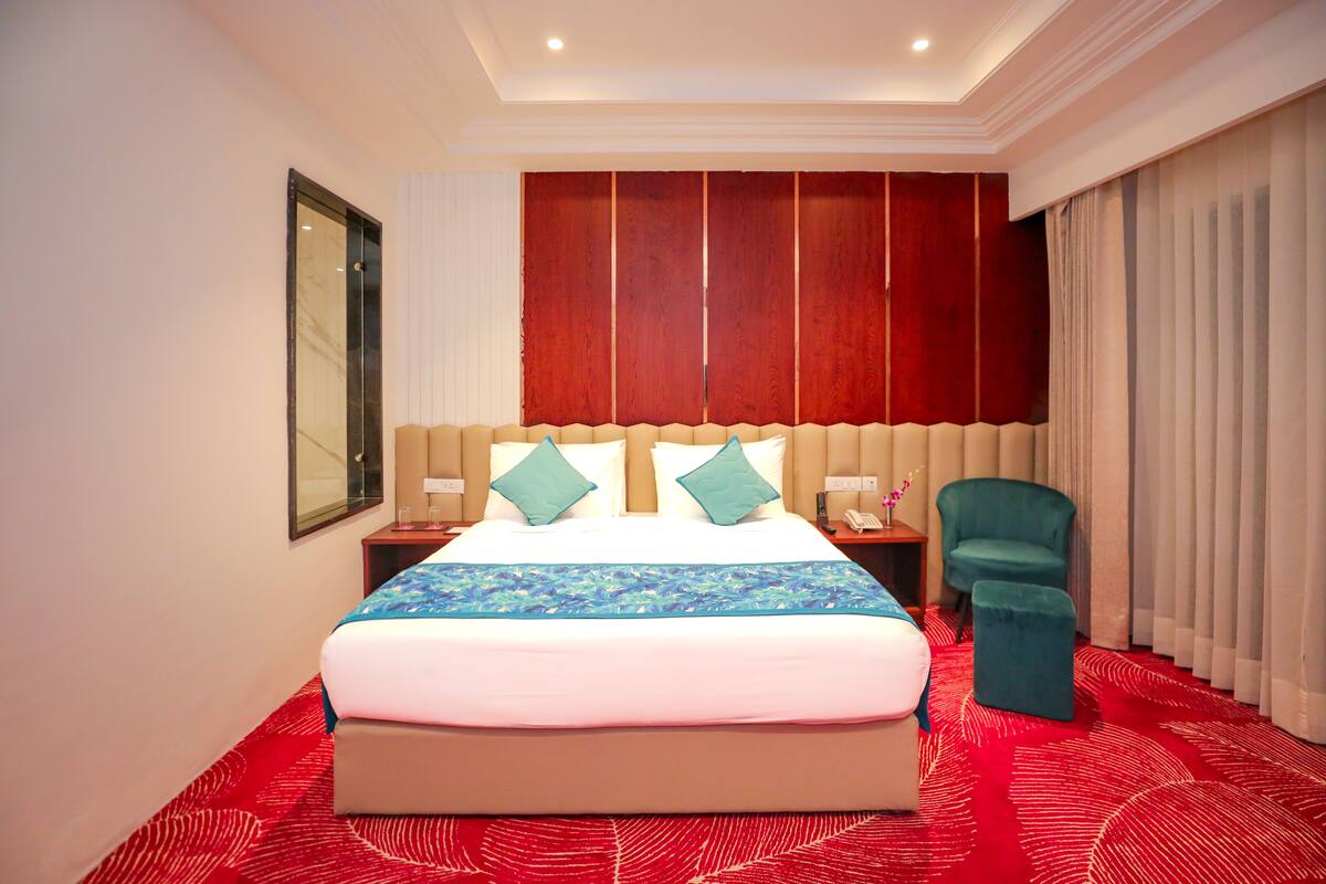 Una habitación de hotel con una cama grande, un cabecero de madera, una alfombra roja, un sillón verde azulado y un ventanal con cortinas.
