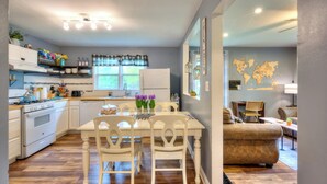 A modern kitchen with white cabinets, a white refrigerator, and a dining area with a white table and chairs. There is a world map on the wall and a window with curtains. - Laurna Dune (Gary)