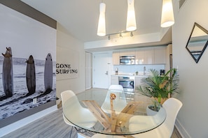 A modern dining area with a glass table, white chairs, and a kitchen in the background.