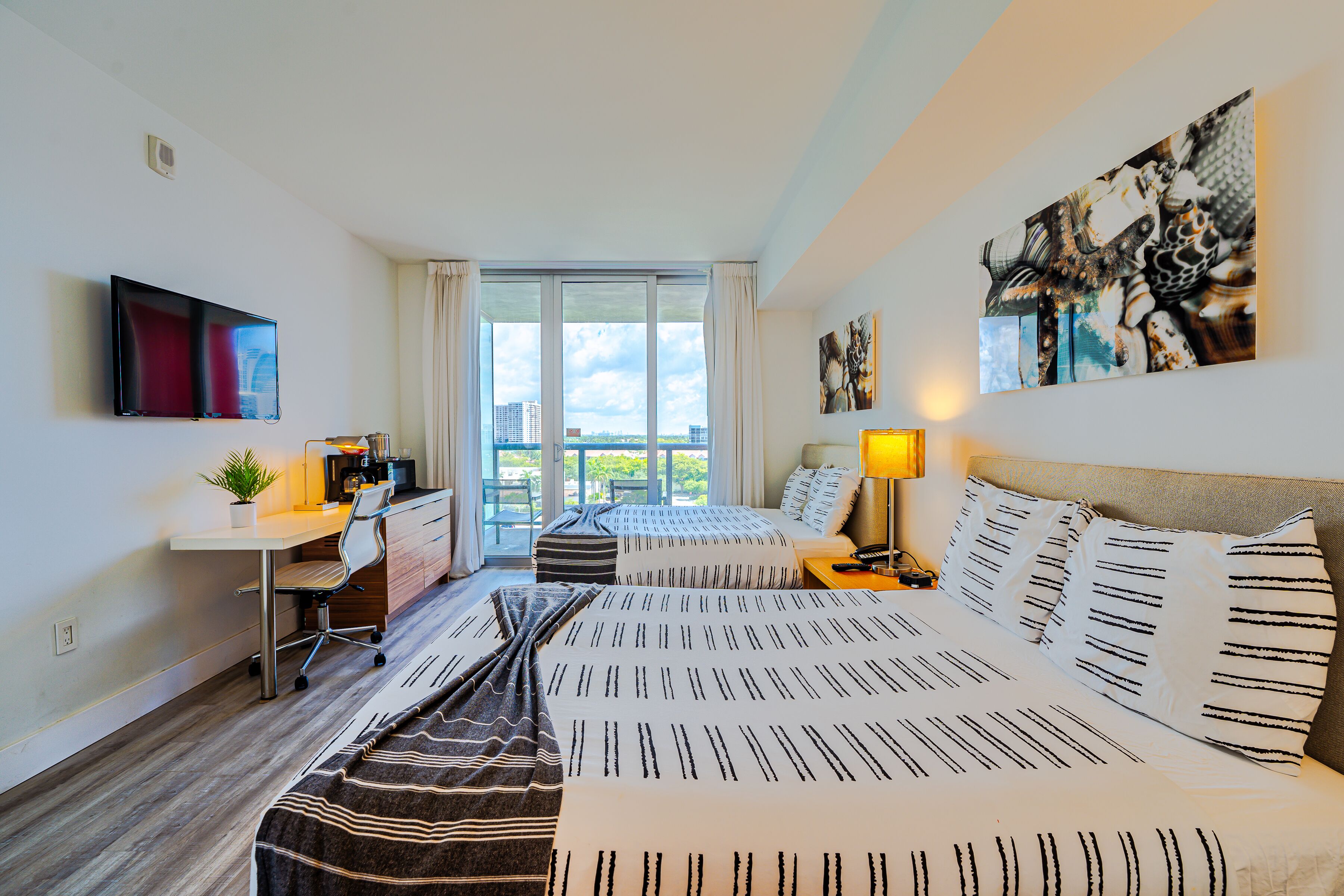 A hotel room with a bed, a desk, a chair, a TV, and a large window with a view of buildings.