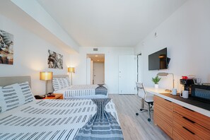 A modern hotel room with two beds, a desk, a TV, and a kitchenette.