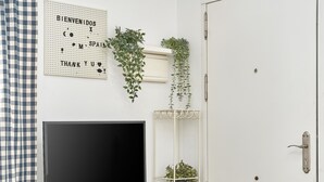 Una habitación con una televisión sobre un soporte, un cartel en la pared con saludos, una puerta y un estante blanco con plantas.