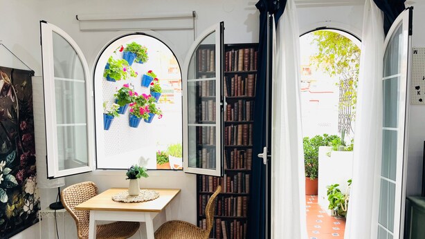 Una habitación con una mesa, sillas, una estantería y una ventana que da a un jardín.