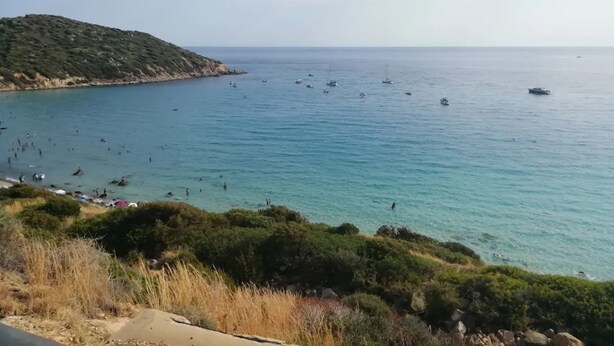 Paisagem costeira com vista clara para o mar, barcos e um terreno montanhoso.