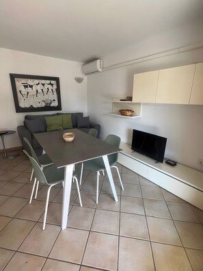 Un salon moderne avec un canapé gris, une table à manger avec des chaises, un téléviseur à écran plat et une étagère murale agrémentée d’un panier.