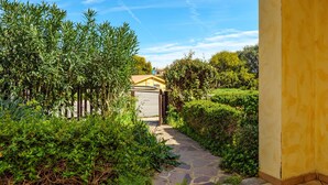 A pathway leading to a building with a yellow exterior, surrounded by lush greenery and a clear blue sky.