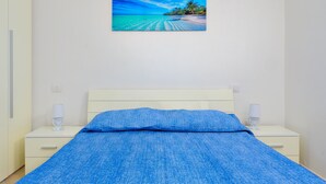 Un dormitorio con una cama, mesitas de noche blancas y un cuadro de una playa.