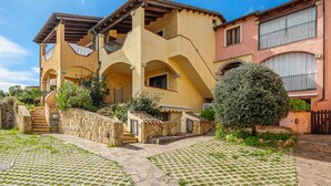 Un edificio de varios pisos con una escalera de piedra que conduce a un balcón, rodeado de follaje y un patio empedrado.
