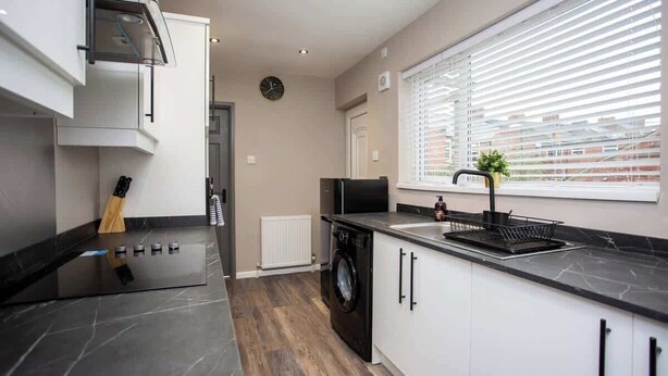 A modern kitchen with a built-in washer, a black countertop, and a window with blinds. - Modern Interior Spacious  Four Bedroom Apartment (South Shields)