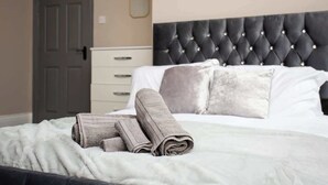 A neatly made bed with rolled towels, a tufted headboard, a white dresser, and a door in a hotel room. - Modern Interior Spacious  Four Bedroom Apartment (South Shields)