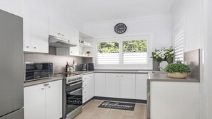Cozinha moderna com armários brancos, forno embutido, micro-ondas e máquina de café. Há um relógio na parede e uma janela com persianas.