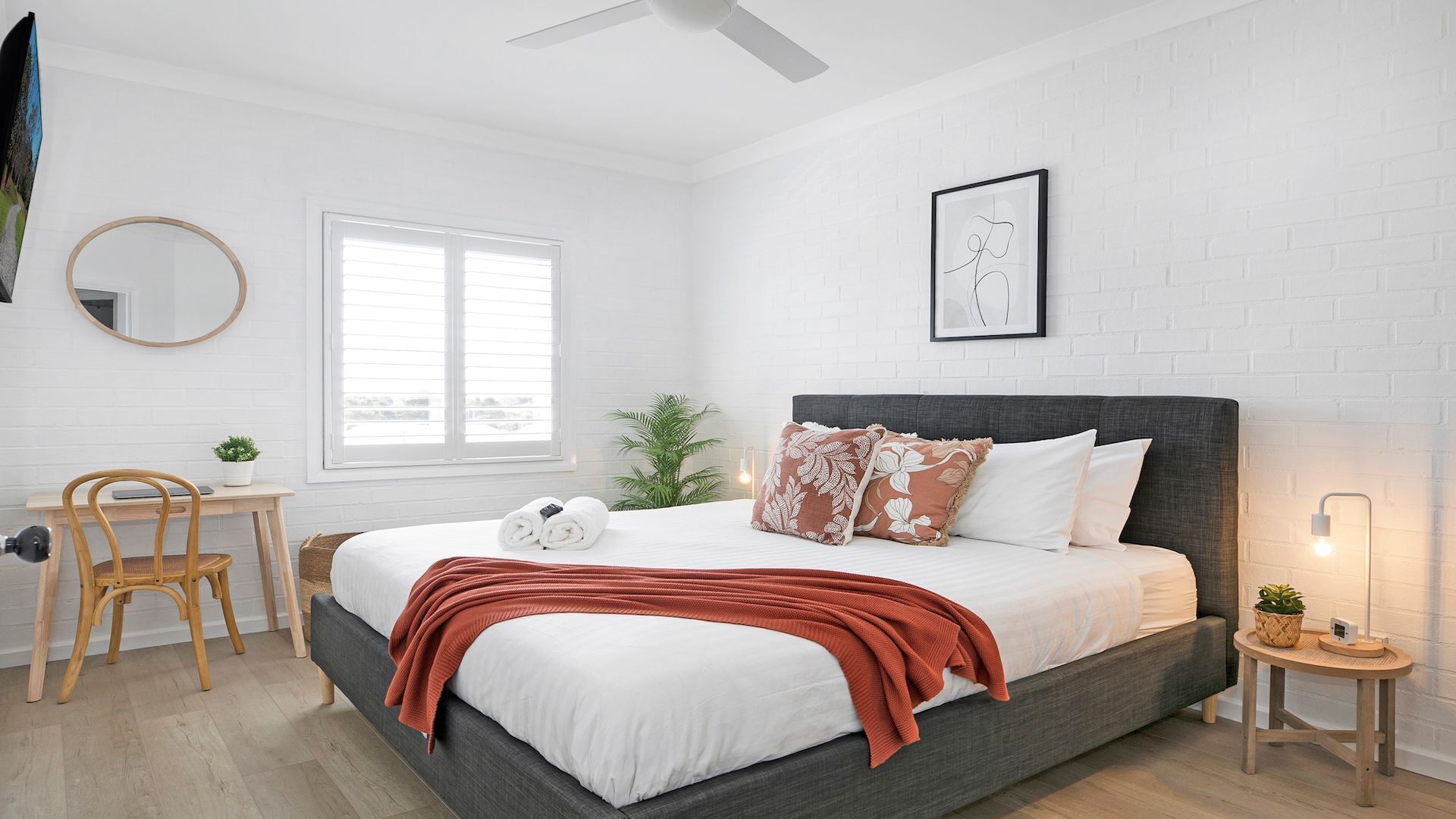 Un dormitorio moderno con cama, escritorio, silla, espejo redondo, ventana y una obra de arte colgada en la pared.