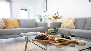 Uma sala de estar moderna com uma mesa de centro de vidro exibindo uma tábua de frios, um sofá cinza e um vaso de plantas.