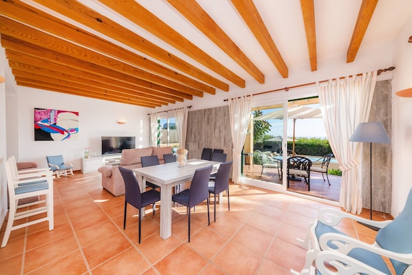 A spacious living area with a dining table, chairs, a sofa, a TV, and a view of the outdoors.
