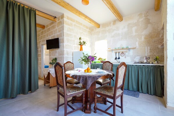 A dining area with a round table, chairs, a TV mounted on the wall, and a kitchen in the background.