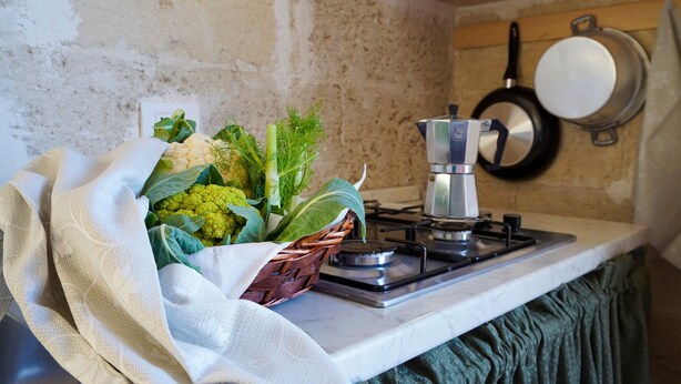 A kitchen with a gas stove, a basket of fresh vegetables, and a hanging pan.