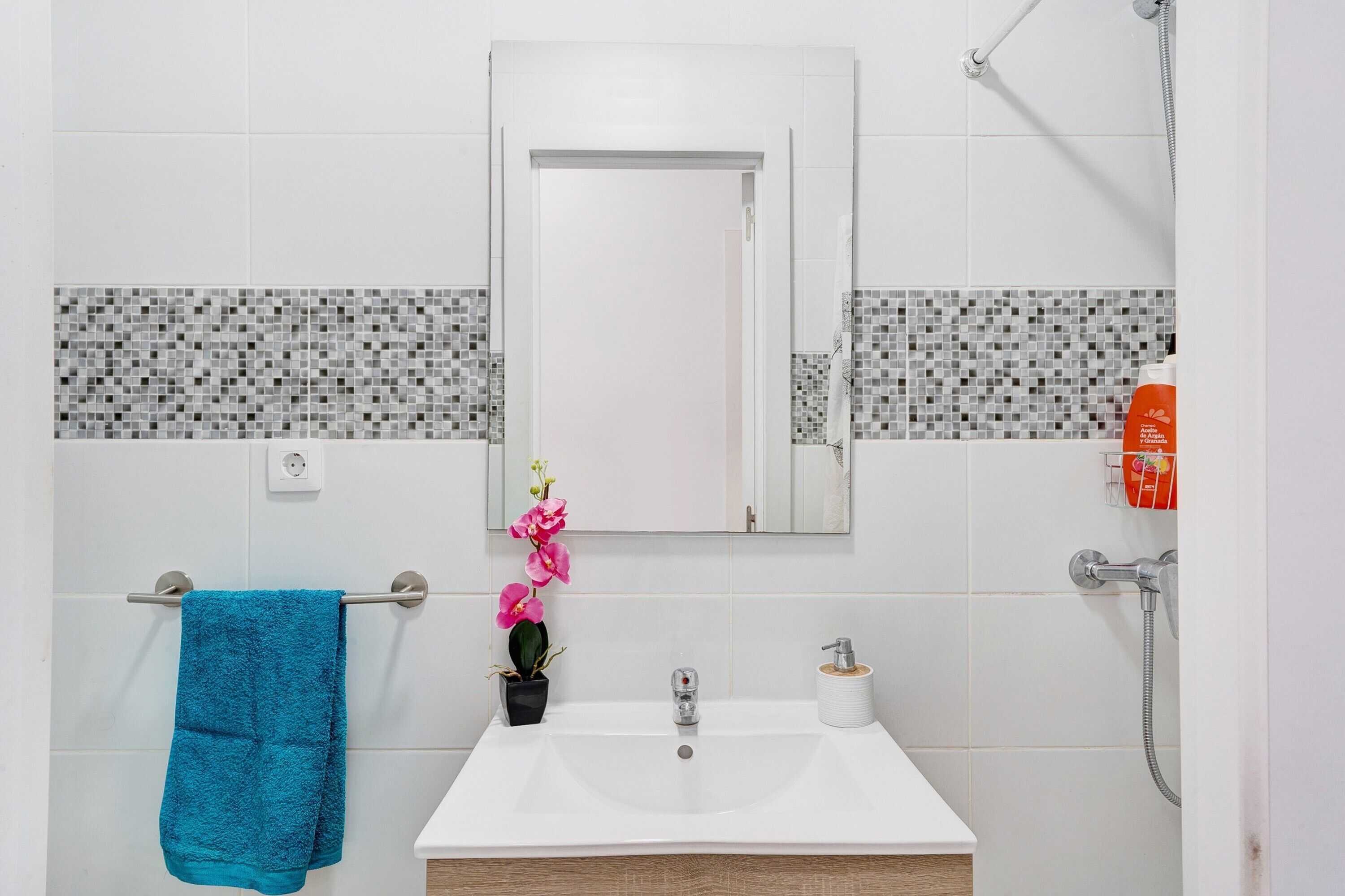 Un baño con un lavamanos blanco, un espejo, un perchero con una toalla azul, una orquídea en maceta y una ducha.