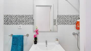 Un baño con un lavamanos blanco, un espejo, un perchero con una toalla azul, una orquídea en maceta y una ducha.