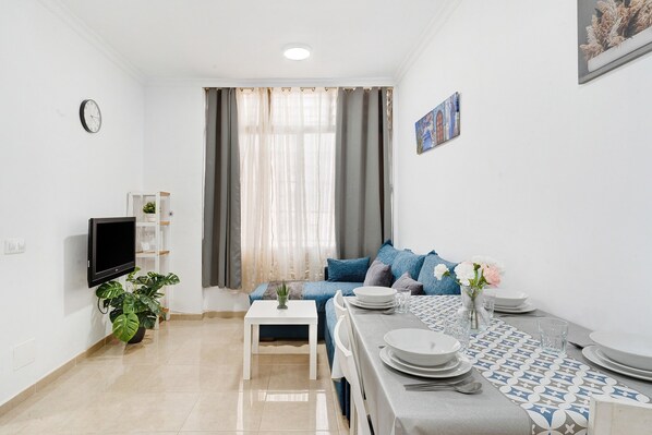 A modern living room with a sofa, dining table set, a TV, and a potted plant.