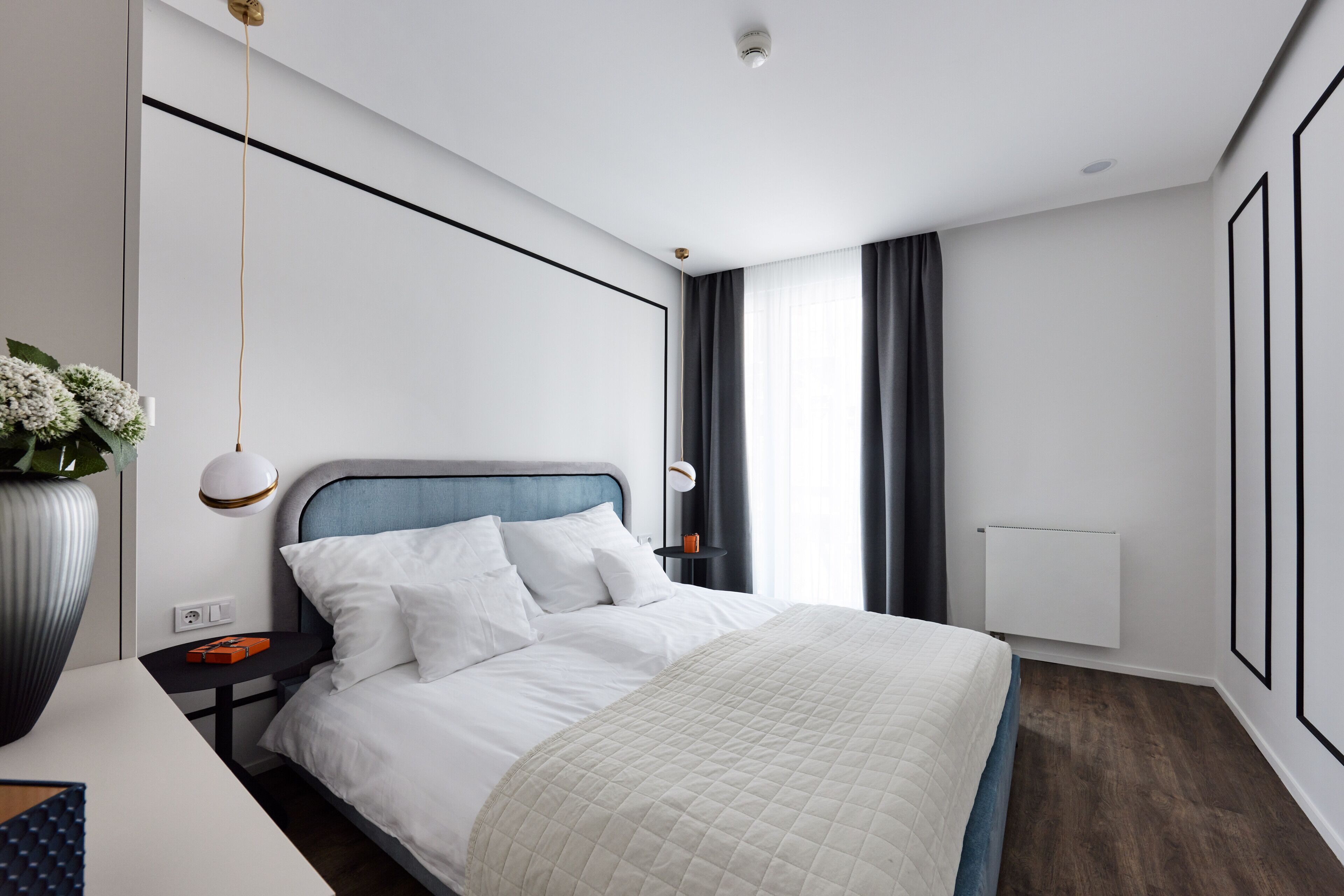 A modern hotel room with a neatly made bed, a bedside table with a lamp, a large vase of flowers, and a view of the outdoors through the window.